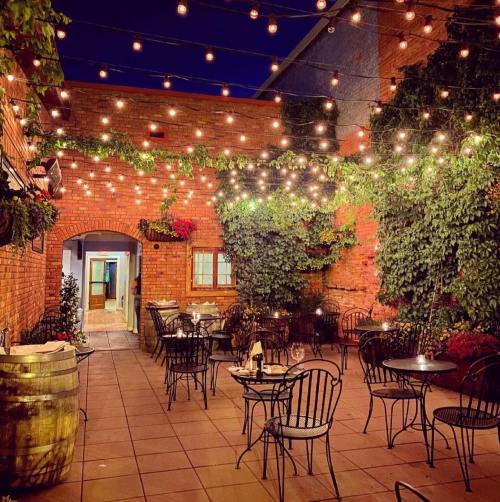 Cozy outdoor dining area with string lights, brick walls, and greenery, featuring tables and chairs set for guests.