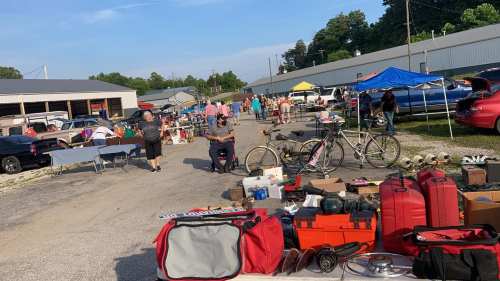 A bustling flea market scene with vendors, shoppers, and various items for sale, including bicycles and luggage.