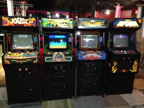 Four vintage arcade game machines lined up in a dimly lit arcade, featuring colorful screens and classic designs.