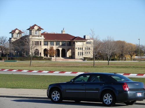 A historic building with a red-tiled roof and a car parked in the foreground, set in a grassy area.