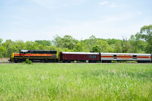 A vintage train with a black locomotive and red passenger cars travels through a green landscape under a clear sky.