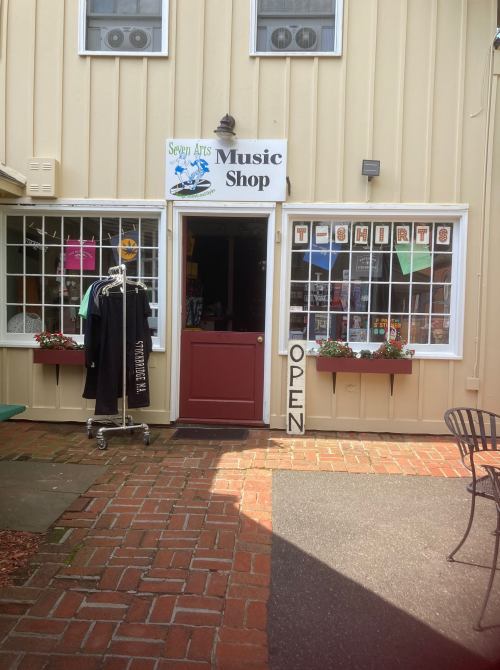 A storefront of a music shop with a sign, open door, and display of T-shirts outside. Brick pathway in front.