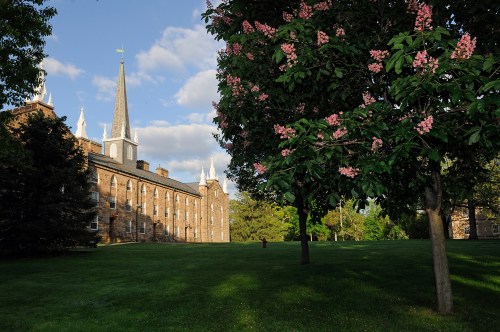A historic building with a tall steeple, surrounded by green grass and blooming trees under a partly cloudy sky.