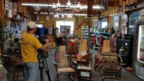 A person films inside a rustic store filled with wooden furniture and various items, while another person sits at a counter.