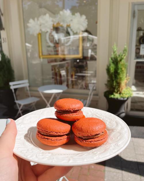 A hand holds a plate with three red macarons in front of a café with outdoor seating.
