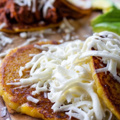 Close-up of golden tortillas topped with shredded cheese, with a side of pulled meat and fresh greens.