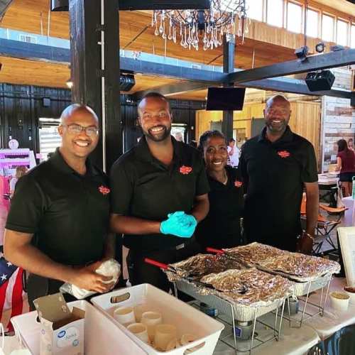Four smiling individuals in black shirts stand behind a food serving station with trays of food in a rustic venue.