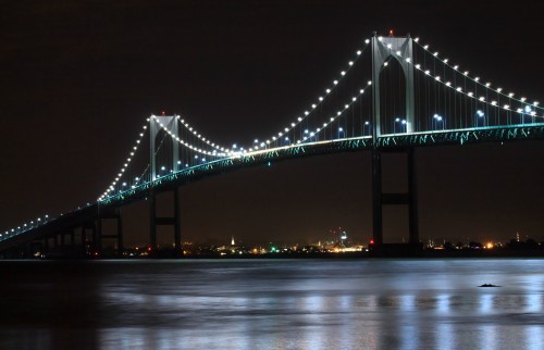 A brightly lit bridge spans over a calm body of water at night, with city lights twinkling in the background.