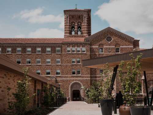 A brick building with a clock tower, surrounded by greenery and a pathway, under a partly cloudy sky.