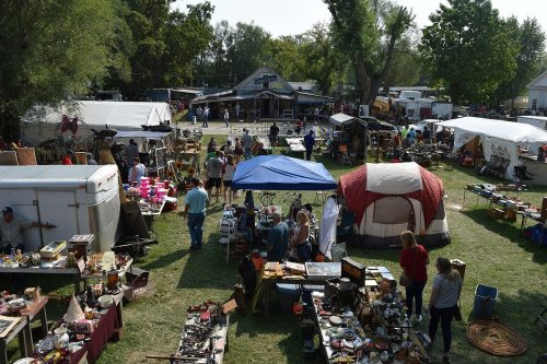 A bustling outdoor market with tents, tables of goods, and people browsing among various items for sale.