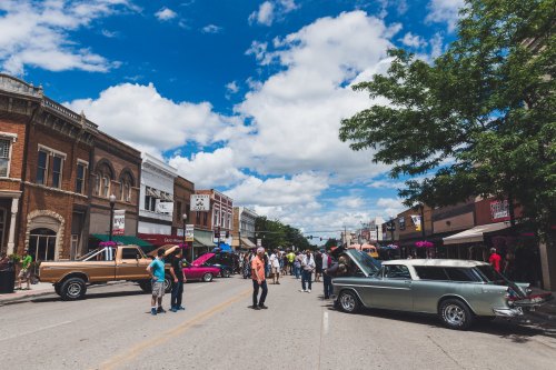 A lively street scene with classic cars on display, people mingling, and a bright blue sky with fluffy clouds.