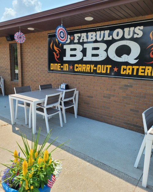 A BBQ restaurant exterior with a sign, outdoor seating, and a colorful flower pot in front.