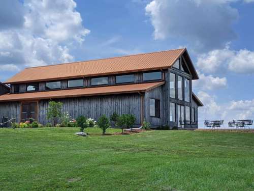 A rustic wooden barn with a metal roof, surrounded by green grass and trees, under a blue sky with fluffy clouds.
