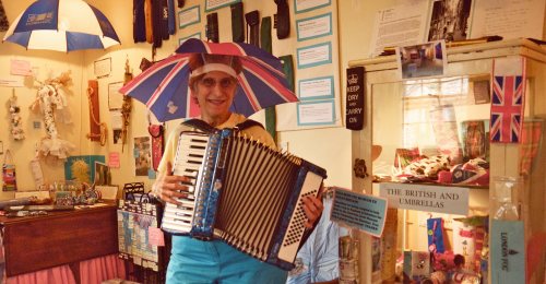 A smiling woman in a colorful umbrella hat plays an accordion in a quirky shop filled with British memorabilia.