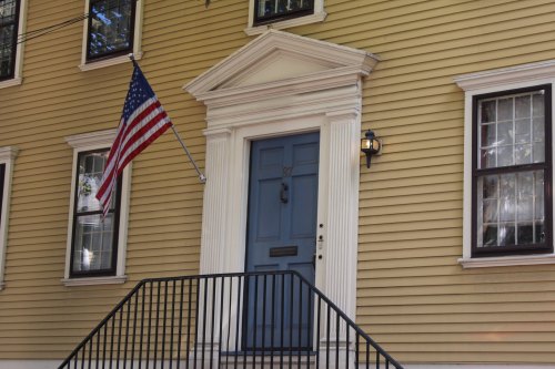 A blue front door with a white frame, flanked by windows, and an American flag hanging beside it on a yellow house.