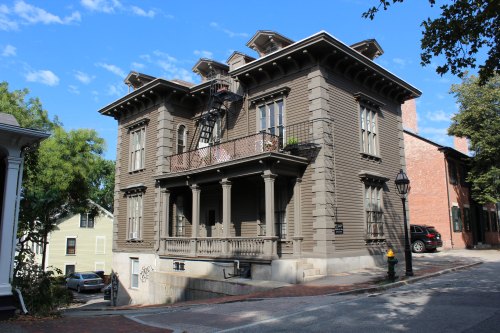 A large, historic brown wooden house with a balcony, set on a sloped street under a blue sky with scattered clouds.