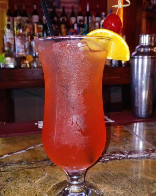 A colorful cocktail in a tall glass, garnished with a cherry and an orange slice, sits on a bar counter.