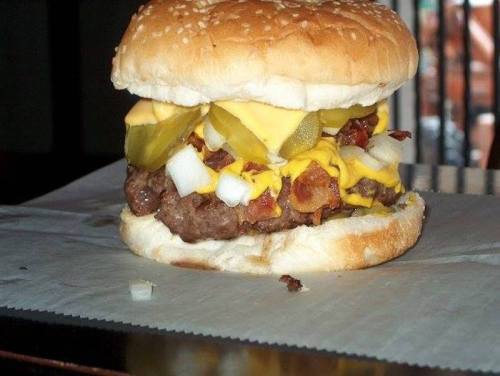 A close-up of a juicy hamburger with cheese, pickles, onions, and bacon on a sesame seed bun, resting on parchment paper.