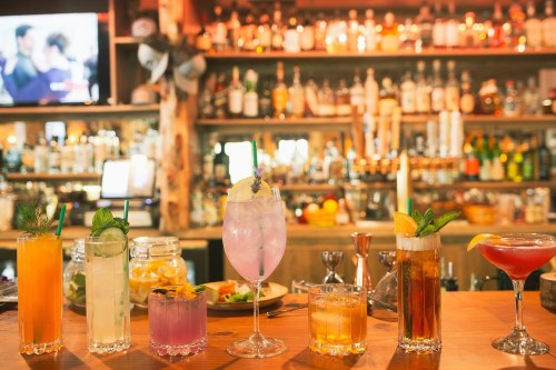 A variety of colorful cocktails lined up on a bar, with a well-stocked shelf of liquor in the background.