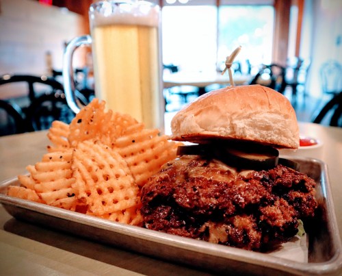 A plate with a juicy burger topped with pickles, served with waffle fries and a glass of beer in the background.
