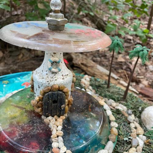 A whimsical garden scene featuring a small, colorful mushroom house surrounded by pebbles and tiny palm trees.