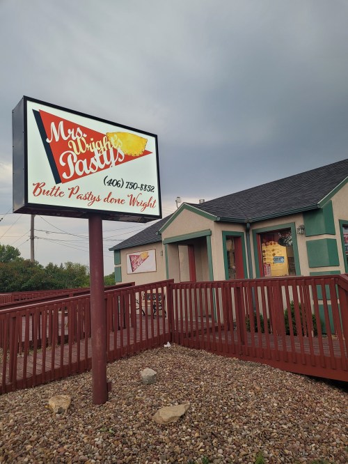 Sign for Mrs. Wright's Pasties, featuring colorful text and a logo, with a building and cloudy sky in the background.