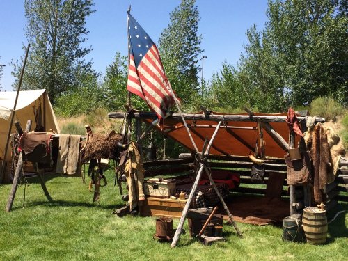 A rustic campsite with an American flag, tents, and various camping gear hanging from wooden structures.