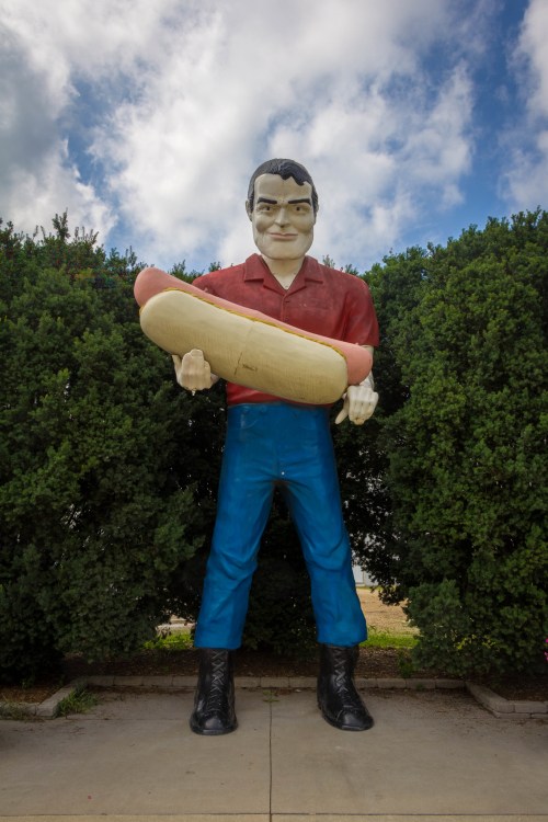 A large statue of a man holding a giant hot dog, standing in front of green bushes under a cloudy sky.