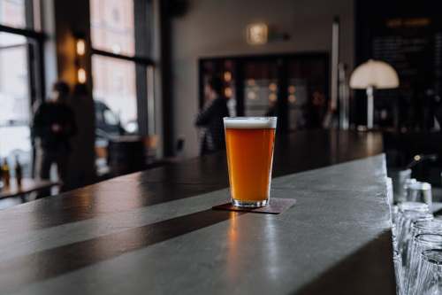 A glass of amber beer sits on a bar counter, with a blurred background of people and a cozy pub atmosphere.