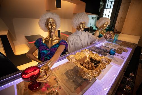 Three golden mannequins with curly white wigs pose at a bar, surrounded by decorative items and colorful glasses.