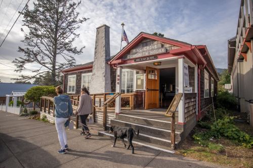 Two people walk a dog past a community house with a wooden exterior and an American flag outside.