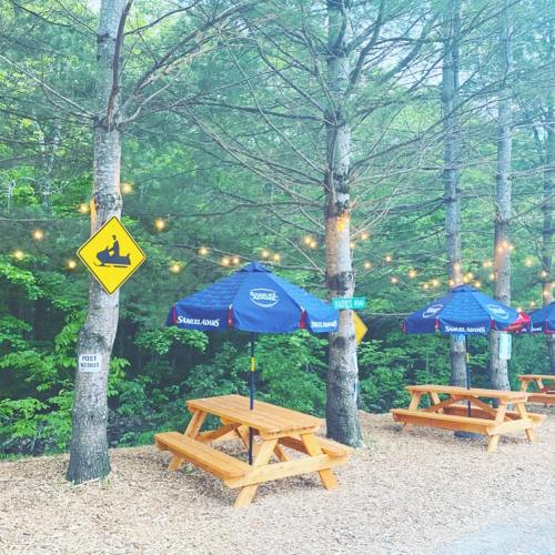 Outdoor seating area with wooden picnic tables, umbrellas, and string lights among trees, with a canoe sign nearby.