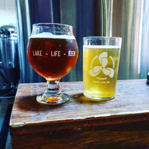Two glasses of beer on a wooden bar: one dark amber and one light yellow, with brewery logos visible.