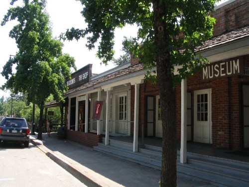 A quaint street scene featuring a brick building with a museum and a shop, surrounded by trees on a sunny day.