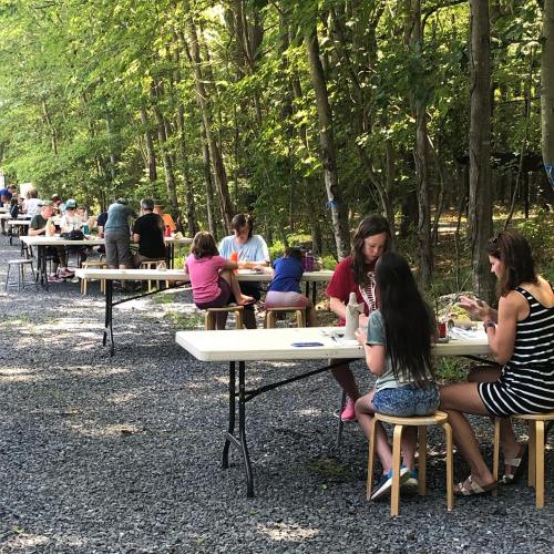 A group of people sitting at tables outdoors, enjoying a meal in a wooded area.