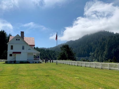 A white house with a red roof, surrounded by green grass and trees, under a blue sky with clouds. An American flag stands nearby.