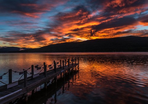 A serene lake at sunset, with vibrant orange and blue skies reflecting on the water and a wooden dock in the foreground.
