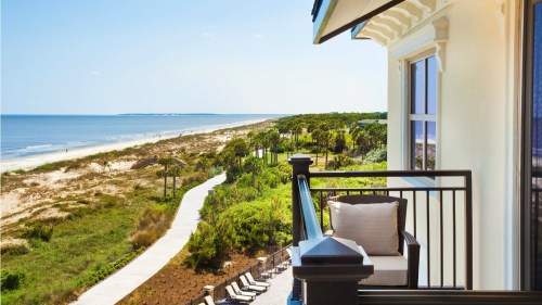 A scenic view from a balcony overlooking a sandy beach, palm trees, and a winding path along the coastline.
