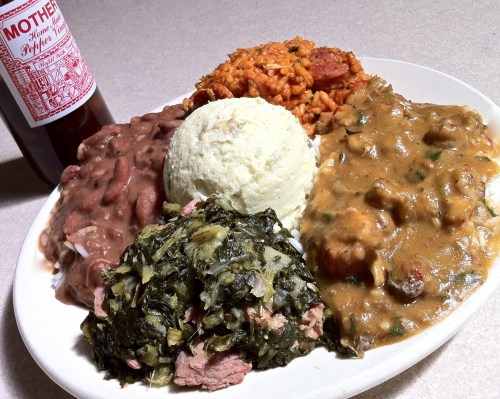 A plate of Southern comfort food featuring rice, beans, greens, mashed potatoes, and gravy, with a bottle of sauce.