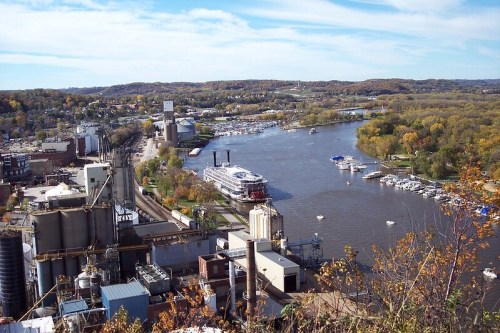 Aerial view of a river with boats, industrial buildings, and autumn foliage along the banks.