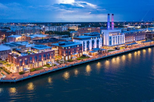Aerial view of a waterfront cityscape at dusk, featuring illuminated buildings along the shore and a calm waterway.