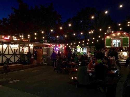 A lively food truck market at night, with string lights illuminating people dining and various food stalls.