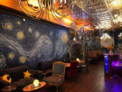 Cozy café interior with a starry night mural, chandeliers, and warm lighting, featuring colorful decor and seating.