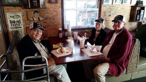 Three elderly individuals sit at a table in a restaurant, enjoying food and drinks, with a cozy, decorated background.