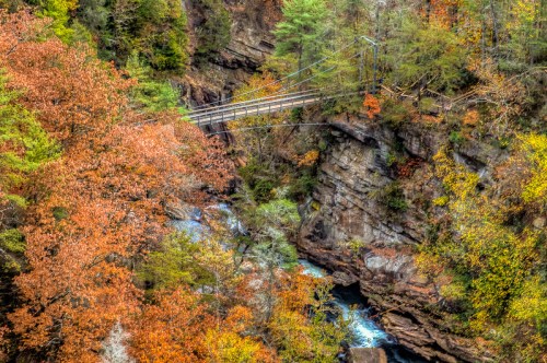 A scenic view of a suspension bridge over a river, surrounded by vibrant autumn foliage and rocky cliffs.