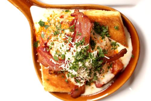 A wooden spoon filled with toast topped with bacon, herbs, and cheese, garnished with spices.
