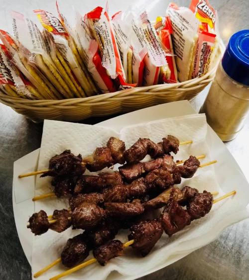 A plate of skewered meat sits on a napkin, with a basket of snacks and a spice container in the background.