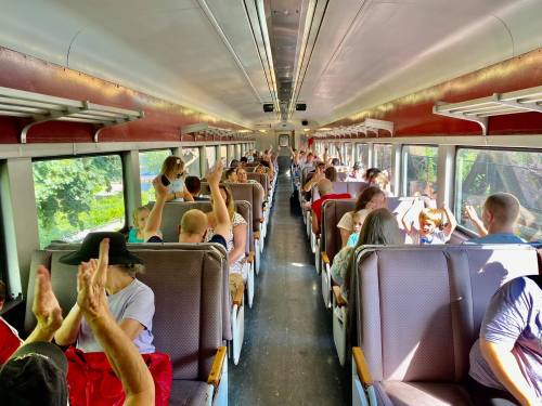 Interior of a train with passengers seated, some raising their hands, enjoying the ride on a sunny day.