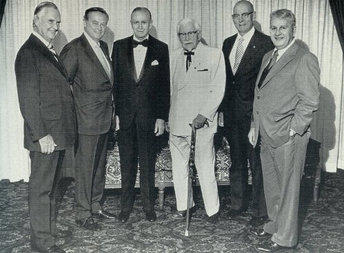 A group of six men in formal attire posing together in a well-decorated room.