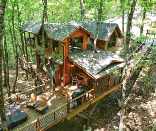 A wooden cabin nestled in a lush forest, featuring large windows and a spacious deck with outdoor seating.